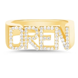 May include: A gold-toned ring featuring the name OREN spelled out in block letters. Each letter is encrusted with small, clear, sparkling stones along the front surface, creating a bright, reflective appearance against the smooth gold-colored metal band.