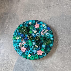 Könnte beinhalten: Ein runder Untersetzer aus Kunstharz mit einer Mischung aus türkisfarbenen, blauen und grünen Perlen. Das Design enthält zwei kleine grüne Froschfiguren, rosa und weiße Blumenanhänger, winzige Pilze sowie zwei dunkelgrüne Blätter als dekorative Elemente.