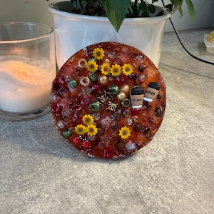 Könnte beinhalten: Ein runder Untersetzer aus Kunstharz mit einem herbstlichen Design, bestehend aus kleinen gelben Sonnenblumen, grünen und roten Perlen, metallischen Akzenten und zwei Miniatur-Kaffeetassen-Anhängern mit dem aufgedruckten Wort COFFEE.
