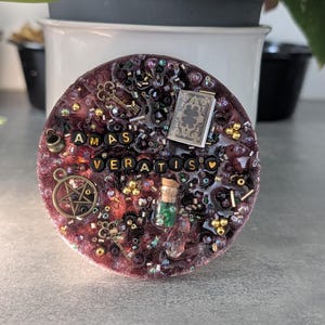 Könnte beinhalten: Ein runder Untersetzer aus dunkelviolettem Harz, verziert mit goldfarbenen Schlüsseln, einem Miniaturbuch, einem Pentagramm-Anhänger, einer kleinen Glasflasche mit Korken und grünem Inhalt sowie schwarzen Buchstabenperlen, die AMAS VERATIS und ein Herz bilden.