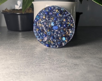 Op de afbeelding: Een ronde onderzetter van hars met een diepblauwe basis ingelegd met diverse blauwe, zilveren en gouden kralen, kleine stervormige lovertjes en twee metalen halve maan bedels, wat zorgt voor een textuurrijk oppervlak met een hemels uiterlijk.