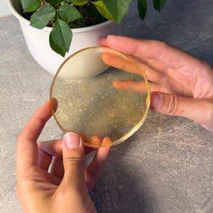May include: A circular, translucent coaster made of resin with a golden, shimmering finish. The coaster has a smooth, flat surface and a clear, slightly amber-toned appearance with fine glitter particles embedded throughout the material.