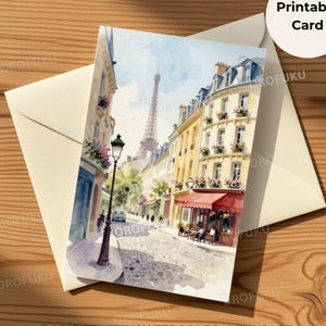 May include: A printable card featuring a watercolor painting of a Parisian street scene with the Eiffel Tower in the background, a black lamppost, cobblestone streets, and buildings with red awnings and flower-filled balconies on a cream envelope.