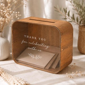 Könnte beinhalten: Eine rechteckige Holzbox mit einer transparenten Frontscheibe, die den weißen Schriftzug THANK YOU for celebrating with us und eine kleine Blattgrafik zeigt. Die Box hat einen Schlitz an der Oberseite und enthält hellbraune Umschläge.