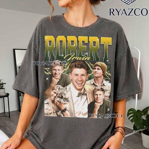 May include: A charcoal gray short-sleeve t-shirt featuring a graphic print of Robert Irwin. The design includes the name "ROBERT" in large yellow and green letters, "Irwin" in script, and four photos of Robert Irwin, one of which shows him holding a koala.