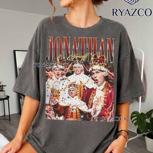 May include: A charcoal grey short-sleeve t-shirt featuring a graphic print of Jonathan Groff in royal attire with crowns and red lightning accents. The text JONATHAN GROFF is printed in a bold, metallic-style font above the central image.