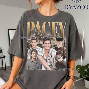 May include: A charcoal gray short-sleeve t-shirt featuring a large graphic print on the front. The design includes the gold text PACEY above a collage of five images of the same person in various outfits, set against a background of palm trees.
