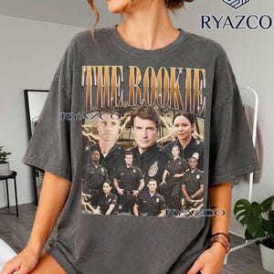 Può includere: Una t-shirt grigio antracite a maniche corte con una stampa grafica del cast di The Rookie. Il design include il testo THE ROOKIE in un carattere oro metallizzato sopra un gruppo di agenti di polizia in uniforme con accenti a forma di fulmine giallo.