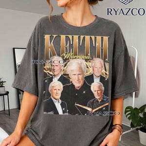 Könnte beinhalten: Ein anthrazitfarbenes Kurzarm-T-Shirt mit einem grafischen Druck von Keith Morrison. Das Design zeigt den Namen KEITH in großen goldenen Buchstaben, den Namen Morrison in Schreibschrift und vier verschiedene Porträtfotos der Person.