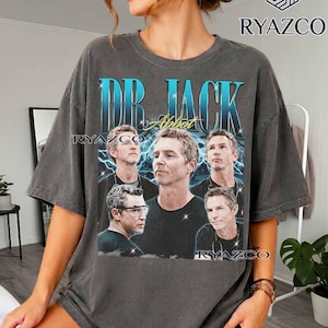 May include: A charcoal grey short-sleeve t-shirt featuring a graphic print with the text DR. JACK and Abbot. The design includes five photographic portraits of a man in various poses against a blue lightning background with sparkling star accents.