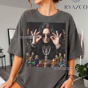 May include: A charcoal gray short-sleeve t-shirt featuring a graphic print of a person with long dark hair wearing round sunglasses and layered silver chain necklaces. The print includes several colorful glass bottles on a table and the text RYAZCO.