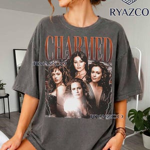May include: A charcoal gray short-sleeve t-shirt featuring a graphic print of four women with the text "CHARMED" in large orange letters above them. The design includes lightning bolt accents and the brand name "RYAZCO" printed in multiple locations.