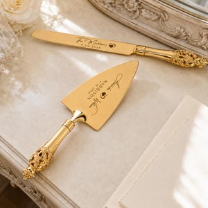 Puede incluir: Un juego de cuchillo y pala para pastel en tono dorado con mangos ornamentados de estilo filigrana. El cuchillo tiene grabado "The Maddison's 9.10.2026" y un corazón. La pala tiene grabado "Amanda & William", "MADDISON" y "9.10.2026" con un corazón.