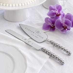 Puede incluir: Un juego de cuchillo y pala para pastel en plata con mangos de cuentas esféricas. La pala está grabada con "EVELYN + COLE" y "10.21.2026". El cuchillo está grabado con "E + C" y "10.21.2026". Ambas piezas tienen un acabado metálico pulido.