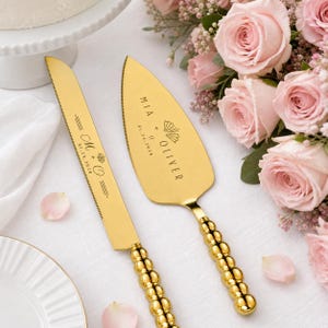 Puede incluir: Un juego de cuchillo y pala para pastel en tono dorado con mangos de cuentas. El cuchillo está grabado con M + O y 01.24.2028. La pala presenta el texto MIA + OLIVER y 01.24.2028 con un icono de abanico. Acabado metálico pulido.