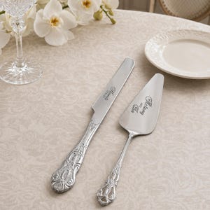Puede incluir: Un juego de cuchillo y pala para pastel en tono plateado con mangos ornamentados y en relieve. La hoja del cuchillo está grabada con el nombre 'Francis' y la fecha '07.01.23', mientras que la pala presenta el texto grabado 'Melany and Leo'.