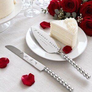 Puede incluir: Un juego de cuchillo y pala para pastel en plata con mangos de cuentas. La pala está grabada con "MARGOT + BLAKE" y "02.22.2028". El cuchillo está grabado con "M + B" y "02.22.2028". Ambas piezas tienen un acabado metálico pulido.