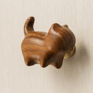 Puede incluir: Un pomo de mueble de madera marrón oscuro con forma de gato y cola erguida. La figura de madera tallada presenta un acabado liso y pulido, y está fijada a la superficie mediante una base de montaje circular de metal color latón.