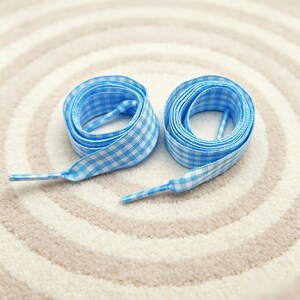 May include: A pair of flat shoelaces featuring a light blue and white gingham checkered pattern. Each lace is coiled into a circular shape with the plastic aglet tip extending outward from the center of the coil, displaying the woven fabric texture.