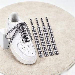 Puede incluir: Una zapatilla blanca con puntera perforada y suela de goma gruesa, con cordones estampados en pata de gallo en blanco y negro. Cuatro cordones adicionales a juego con el mismo estampado están dispuestos en fila junto a la zapatilla.