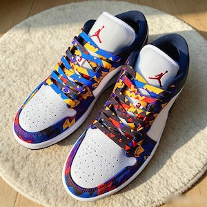 May include: A pair of low-top trainers featuring white perforated leather toe boxes, dark blue collars, and white tongues with red Jumpman logos. The laces and overlays display a vibrant geometric pattern in blue, yellow, red, and black.