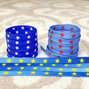 May include: Three sets of flat shoelaces. One coiled set is royal blue with white stars. Another coiled set is light blue with red six-pointed stars. The third set is laid flat, featuring a gradient blue background with yellow stars.