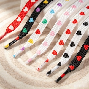 May include: A collection of flat shoelaces featuring repeating heart patterns in various colors. The set includes red, black, and white laces with hearts in shades of red, yellow, blue, green, purple, and pink, each finished with plastic aglets.