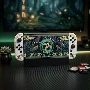 May include: A white handheld gaming console featuring a black screen with a green and gold graphic design depicting a central emblem, stylised figures, and ancient symbols. The console is held in a black stand with small game cartridges nearby.