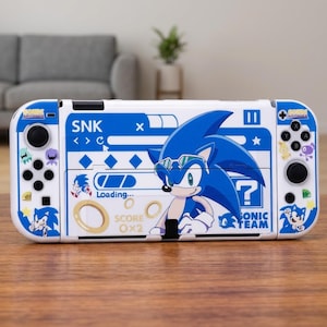 Puede incluir: Una consola de juegos portátil blanca con una funda gráfica azul de Sonic the Hedgehog. El diseño incluye el texto SNK, Sonic Team y Loading, junto con varios iconos temáticos de juegos como anillos, un signo de interrogación e ilustraciones.