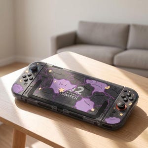 May include: A Nintendo Switch console featuring a black protective case decorated with purple blob-shaped characters, small yellow stars, and crescent moons. The design includes a glittery finish over the black background and the Nintendo Switch logo.