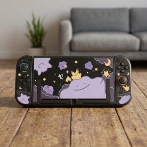Puede incluir: Una consola de juegos portátil negra con una cubierta personalizada que muestra un fondo oscuro, personajes morados en forma de mancha, una criatura amarilla y estrellas amarillas. Incluye mandos negros con botones y joysticks.
