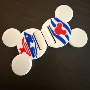 May include: A white plastic magnetic clip shaped like two connected Mickey Mouse heads. The clip features a blue and white striped pattern with a red Mickey Mouse icon on one side and a red and blue graphic design on the other side.