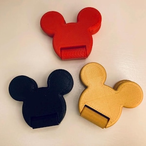 May include: Three plastic phone stands shaped like a mouse head, each featuring a circular top and two rounded ears. The set includes one red, one black, and one gold stand, all displaying a textured surface finish from the manufacturing process.