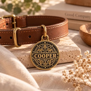 Könnte beinhalten: Ein braunes Lederhalsband für Haustiere mit Messingbeschlägen und einer runden Messingmarke in Antik-Optik. Die Marke zeigt ein verziertes, symmetrisches Blumenmuster, das den Namen COOPER in Großbuchstaben umrahmt, mit kleinen Sternen am Rand.