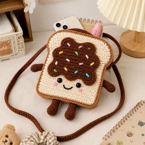 Pode incluir: Uma bolsa transversal de crochê em formato de fatia de pão torrado com cobertura de chocolate marrom, granulados coloridos e um rosto sorridente. A bolsa possui pequenos braços e pernas marrons, uma borda marrom escura e uma alça longa de crochê.