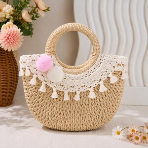 May include: A woven straw handbag featuring a circular handle, a wide white lace trim along the top edge, and a row of small white tassels. Two soft pom-poms, one light pink and one white, are attached to the front of the lace detailing.