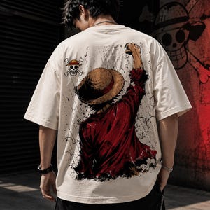 Könnte beinhalten: Ein cremefarbenes Kurzarm-T-Shirt mit einem großen Grafikdruck auf der Rückseite. Das Motiv zeigt eine Figur mit Strohhut und rotem Hemd, die eine Faust in die Höhe streckt, sowie ein kleines Totenkopf-Symbol mit gekreuzten Knochen.