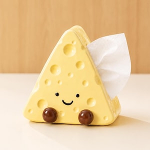 以下が含まれることがあります： 円形のくぼみがあるスイスチーズの形をした、三角形の黄色い陶器製ナプキンホルダー。前面には、2つの小さな目と曲線的な口が描かれたシンプルな黒い顔があり、底面には2つの濃い茶色の球形の足が付いています。