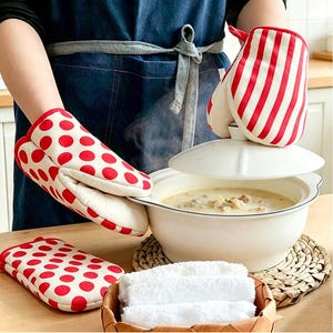 May include: A set of oven mitts featuring a red polka dot pattern on a cream background and a red and white striped pattern. The mitts are shown in use with a white ceramic pot containing a creamy soup, with an additional matching mitt nearby.