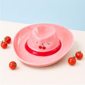May include: A light pink ceramic bowl shaped like a cowboy hat with a wide brim and a raised center. The bowl features a bright red band around the base of the crown and a small painted cherry graphic on the front. Four small red tomatoes are nearby.