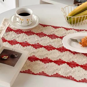 May include: A rectangular woven table runner featuring a repeating pattern of cream-colored diamond shapes and bold red horizontal stripes with fringed edges. The runner is displayed on a white surface alongside a ceramic cup, a plate, and a book.