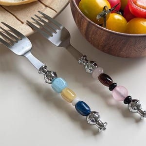May include: Two stainless steel forks with decorative handles featuring colorful beads. One handle has light blue, yellow, white, and dark blue beads. The other handle has pale pink, dark brown, black, and light pink beads, with silver accents.