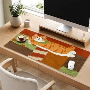 May include: A rectangular desk mat featuring a stylised illustration of an orange tabby cat with a green collar, lying across a background of textured blocks in shades of brown, orange, and green. The cat has white paws and a simple face.