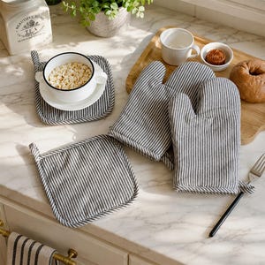 Op de afbeelding: Een set keukentextiel met een donkergrijs en wit krijtstreepmotief, bestaande uit twee vierkante pannenlappen en één ovenwant. De items zijn gemaakt van stof en vertonen een consistent gestreept ontwerp in de gehele collectie.