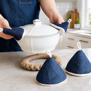 Peut inclure: Une cocotte en céramique blanche avec un couvercle à bouton carré, tenue par deux poignées en jean bleu foncé à bordure blanche. Deux autres poignées coniques en jean avec des boucles blanches et un dessous de plat en jute tressé.