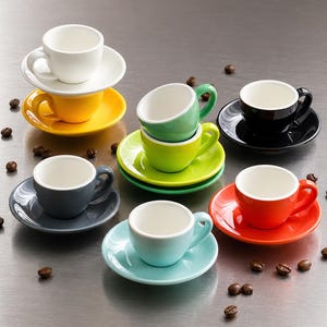 Könnte beinhalten: Eine Sammlung von acht kleinen Espressotassen mit passenden Untertassen in verschiedenen Farben, darunter Weiß, Gelb, Hellgrün, Limettengrün, Schwarz, Dunkelgrau, Hellblau und Orange, arrangiert mit verstreuten gerösteten Kaffeebohnen.