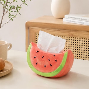 May include: A ceramic tissue box cover shaped like a slice of watermelon. The exterior features a bright red centre with black teardrop-shaped seeds and a thick light green rind along the curved bottom edge. A white tissue is pulled from the top.