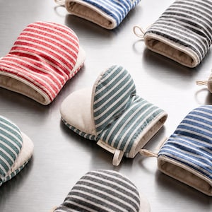 Peut inclure: Une collection de gants de cuisine présentant divers motifs rayés en rouge, bleu, vert et gris. Chaque gant possède une doublure intérieure beige clair et une petite boucle en tissu pour le suspendre. Le texte SEMI CONCEPT est sur une étiquette.