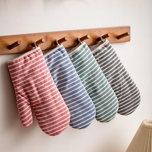 May include: Four oven mitts hanging on a wooden rack. Each mitt features a white and colored striped pattern in red, blue, green, and grey. The mitts are made of fabric and include a small loop at the top for hanging on the wooden pegs.