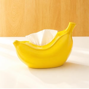 Puede incluir: Un soporte para pañuelos de color amarillo brillante con forma de plátano y acabado liso y brillante. El recipiente de cerámica curvado presenta una abertura superior por la que asoma un pañuelo blanco, mostrando una silueta de fruta realista.